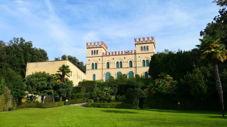 A large, well-maintained garden in front of a castle-like building with a cream-colored facade and battlement-style parapets. The garden features lush green grass, neatly trimmed hedges, and a variety of trees including palm trees and dense shrubs. The soil areas appear moist and dark, indicative of recent maintenance or watering. A gravel pathway runs through the garden, leading towards the building, while small round lights are positioned along the edges of the lawn. The overall scene is illuminated by bright, natural daylight with a clear blue sky overhead, creating a vibrant and inviting outdoor space. This landscape demonstrates careful gardening work with neatly shaped borders and healthy vegetation, typical of garden maintenance and landscaping undertaken by gardenersselephantandcastle.org.uk.